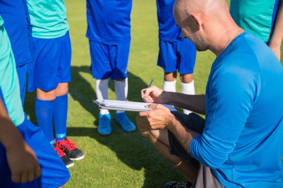 Youth soccer coach kneeling on the grass writing on a clipboard beside young players, with only the players' legs and shorts in frame